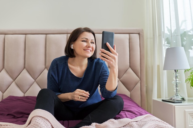 Smiling middle aged woman wearing wireless headphones looking at smartphone