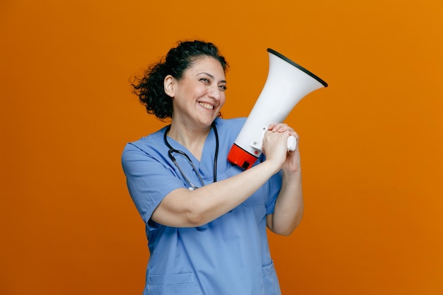 smiling middleaged female doctor wearing uniform and stethoscope around her neck holding speaker with both hands looking at camera isolated on orange background