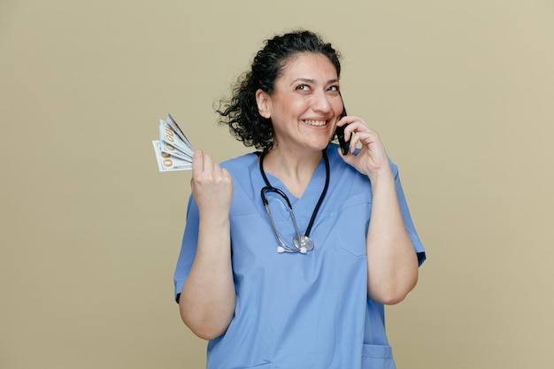 Smiling middleaged female doctor wearing uniform and stethoscope around neck holding money looking up while talking on phone isolated on olive background