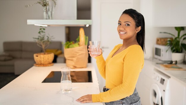 Smiling millennial african american lady in casual holding glass of water in white modern kitchen interior