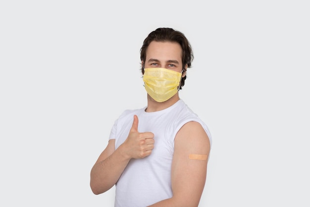 Smiling millennial caucasian man in protective mask show shoulder with band aid after immunization thumb up