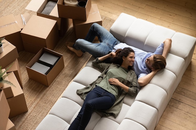 Smiling millennial family couple resting on cozy couch renewing energy after moving cardboard boxes in new apartment planning future decorations or discussing interior ideas together at home