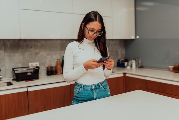 Smiling millennial girl stand at white modern kitchen counter texting messaging on modern cell gadget happy young woman renter using smartphone device with wireless Internet enjoy weekend at home