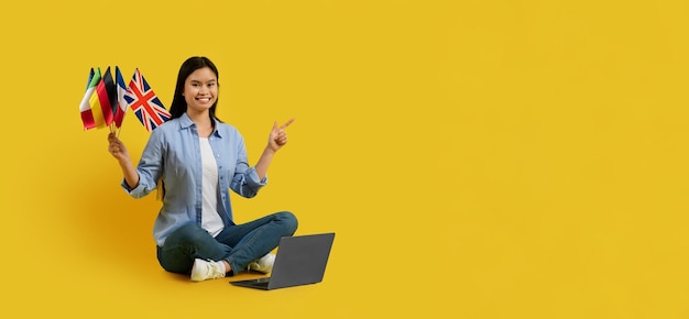 Smiling millennial japanese lady student hold many flags point finger at empty space sit on floor with laptop