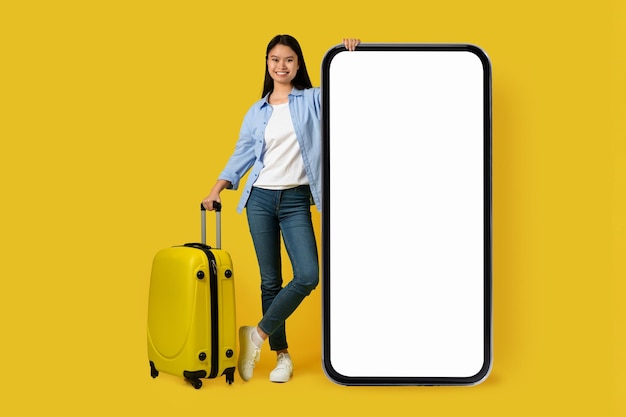 Smiling millennial korean woman student tourist in casual with suitcase stands with huge smartphone