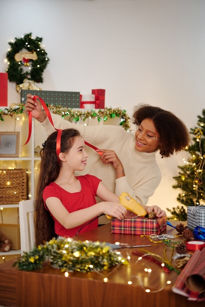 Smiling mixed-race girl tying hair of her friend with red ribbon when they are wrapping presents for Christmas