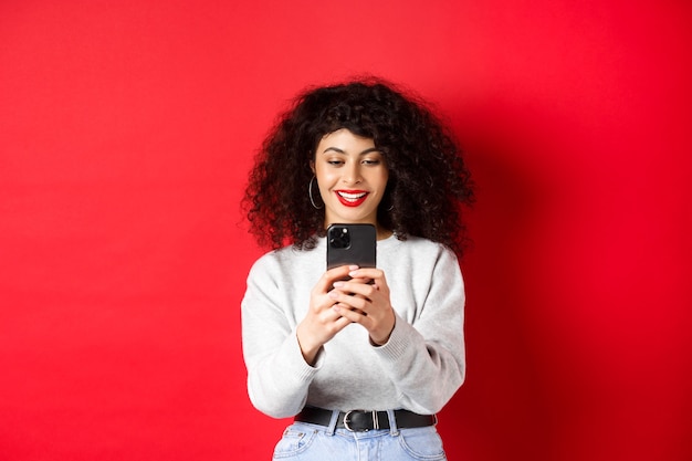 Smiling modern girl taking photos on smartphone, looking at screen and recording video, standing against red wall.