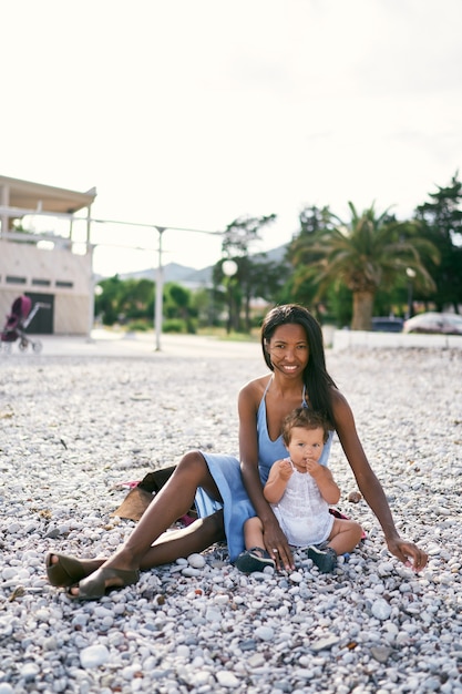 Smiling mom sits on the beach near a little girl gnawing a pebble