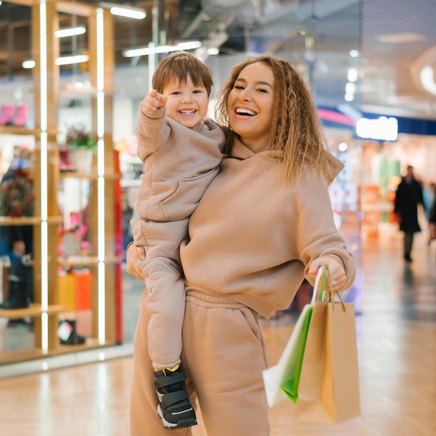 Smiling mother carries her little happy son in her arms holds shopping bags in her hands and walks around the store on sale