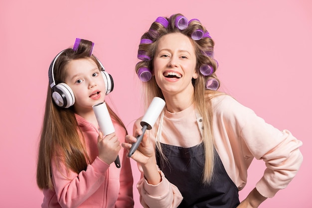 Smiling mother and daughter listen to music sing to a clothes roll have twisted rollers in their hair pose against a pink background