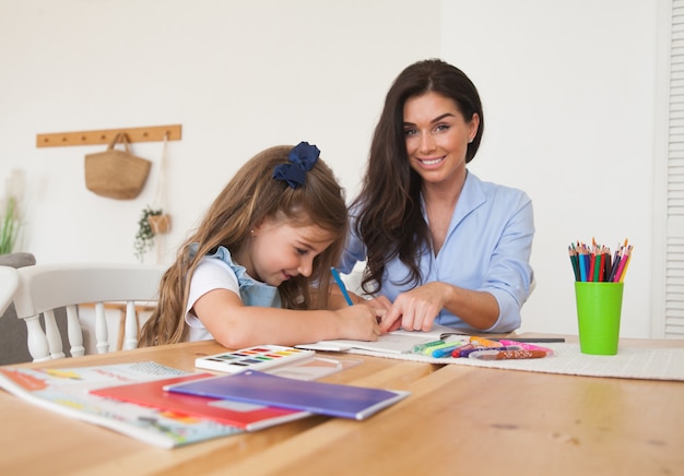 Smiling mother and daughter preparing for lessons and draws at the table with pencils and paints