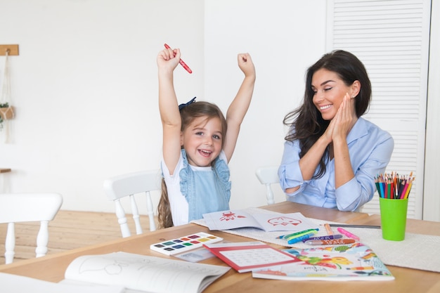 Smiling mother and daughter preparing for lessons and draws at the table with pencils and paints