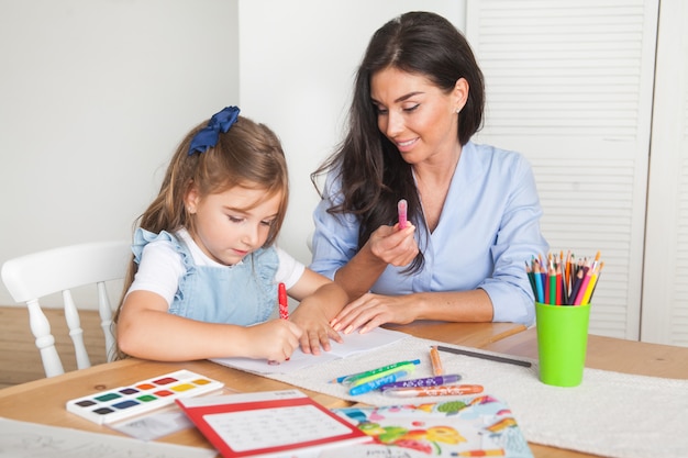 Smiling mother and daughter preparing for lessons and draws at the table with pencils and paints