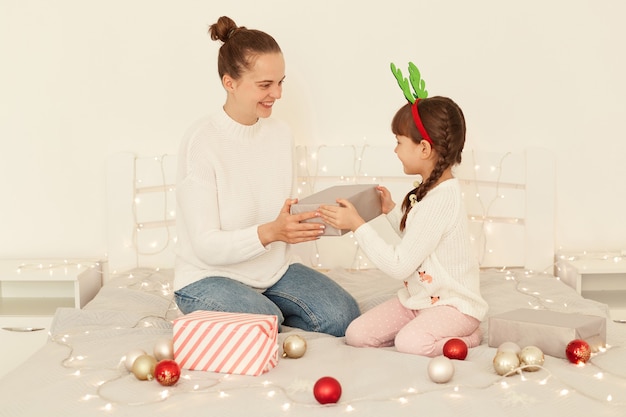 Smiling mother and daughter wearing casual style white sweaters sitting on bed, woman giving present box to her child, congratulating little girl with Christmas and new year.