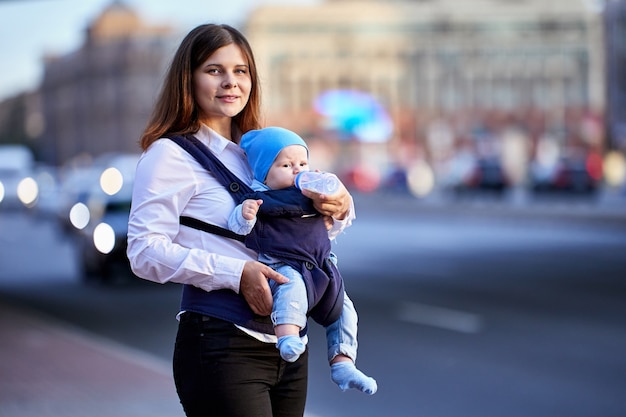 Smiling mother is feeding baby in sling using bottle near traffic