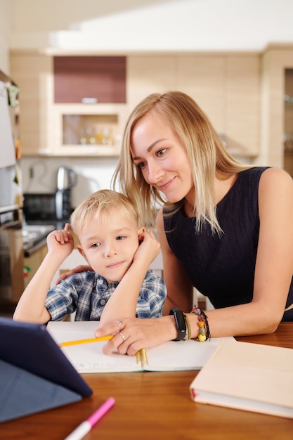 Smiling mother pointing at tablet computer when explaining lesson to her child who is studying at home 