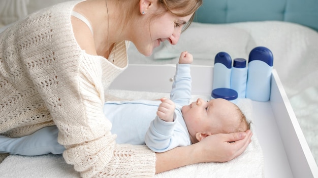 Smiling mother tickling and playing with her little baby son lying on dressing table at bedroom