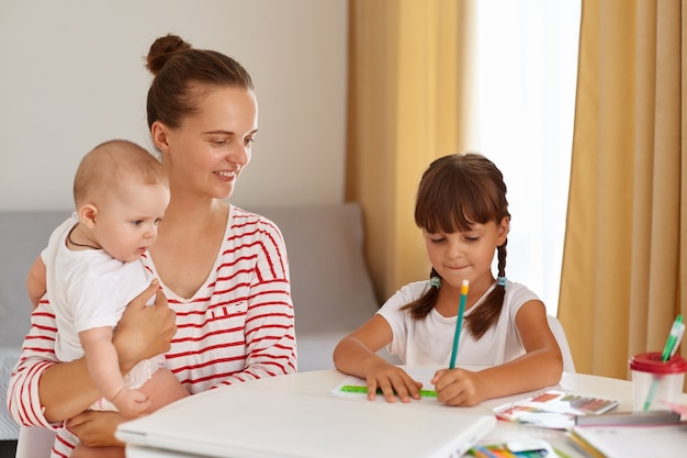 Smiling mother with new born baby in hands and elder daughter sitting at table and doing homework, dark haired girl in white t shirt writing in exercises book or drawing.