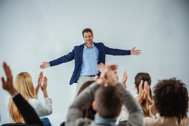 Smiling motivational speaker standing in front of his audience who is clapping.