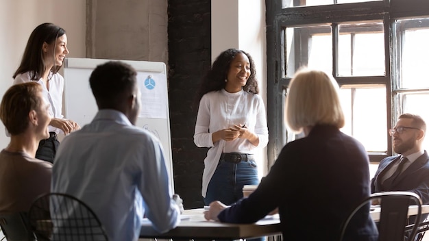 Photo smiling multicultural diverse colleagues brainstorm discuss financial ideas at meeting in boardroom happy multiethnic coworkers employees talk consider flip chart presentation at briefing together