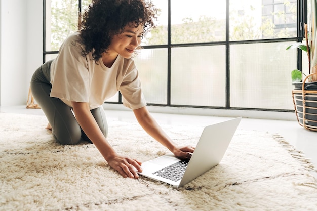 Smiling multiracial latina woman in sports clothes searching fitness video tutorial with laptop to exercise at home Copy space Technology and wellness concept