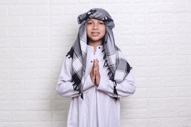 Smiling muslim kid wearing white rob and skullcap is standing and gesturing apologize with hands