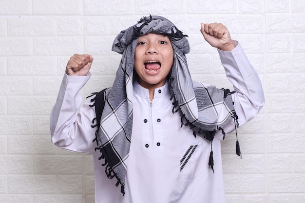 Smiling muslim kid wearing white rob and turban showing celebrating victory while shouting