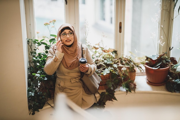 Smiling Muslim woman wearing a hijab and talking on her smartphone while standing by the window.