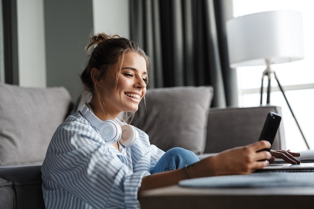 smiling nice woman with headphones using cellphone and laptop while sitting on floor at living room