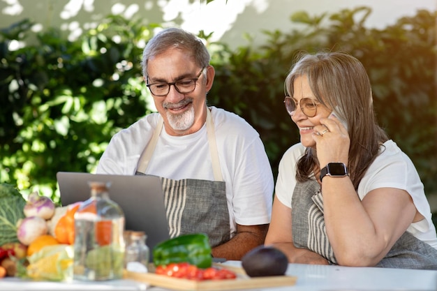 Smiling old caucasian husband and wife in aprons use laptop make phone calls at table with organic