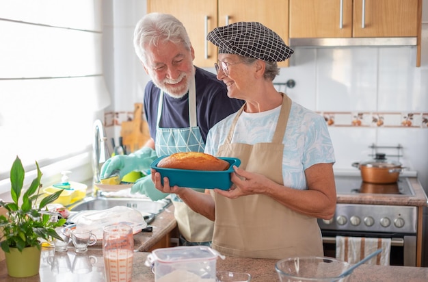 Smiling older woman showing her freshly baked homemade plumcake while her husband washes the dishes Concept of family and domestic partnership