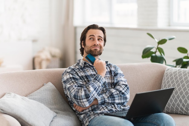 Smiling pensive millennial caucasian man in domestic clothes with stubble dreams of new buy sit on sofa