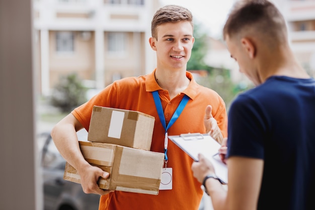 A smiling person wearing an orange T-shirt and a name tag is delivering a parcel to a client, who is putting his signature on the receipt. Friendly worker, high quality delivery service.