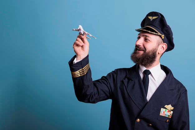 Smiling pilot in uniform holding airplane model, aviation academy aviator playing with plane. Aircraft crew member looking at commercial jet toy, side view studio medium shot