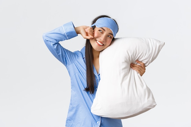 Smiling pleased asian girl in sleeping mask and pyjamas lying in bed and hugging pillow stretching a...