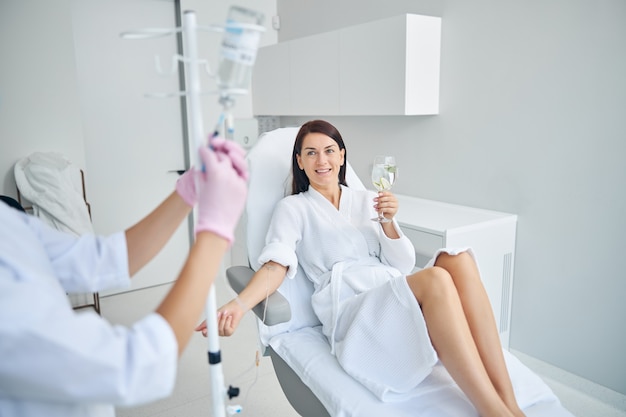 Smiling pleased female patient with a glass of healthy drink sitting in a cosmetic chair