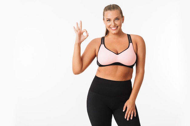 Smiling plump girl in sporty top and leggings happily showing ok gesture while looking in camera over white background