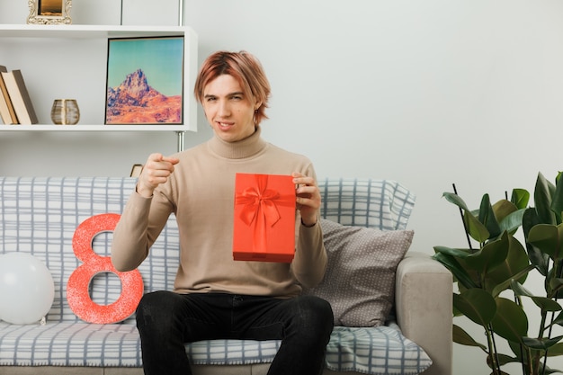 Smiling points at front handsome guy on happy women day holding present sitting on sofa in living room