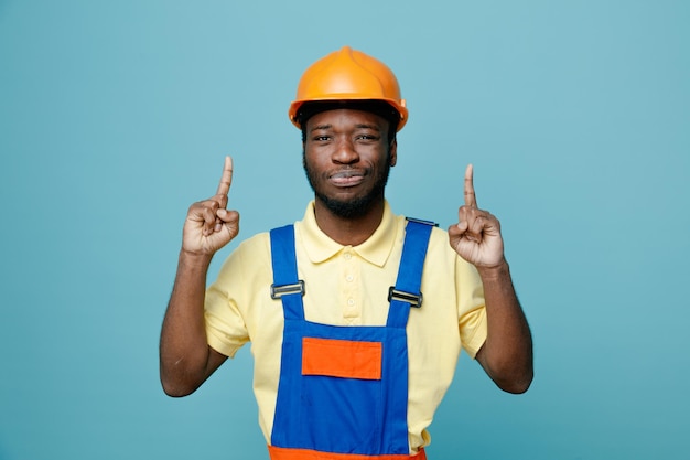 Smiling points at up young african american builder in uniform isolated on blue background