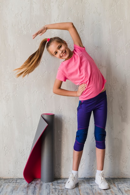 Smiling portrait of a girl doing stretching exercise in front of concrete wall