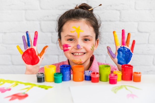 Smiling portrait of a girl behind the table with paint bottles showing her hand and face painted with colors