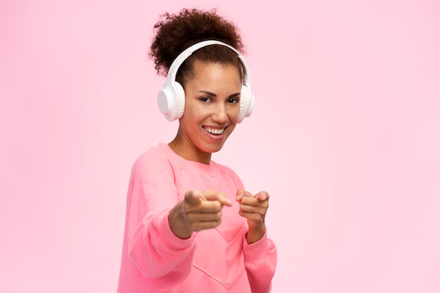 Smiling positive African American woman in earphones points at camera isolated on pink background