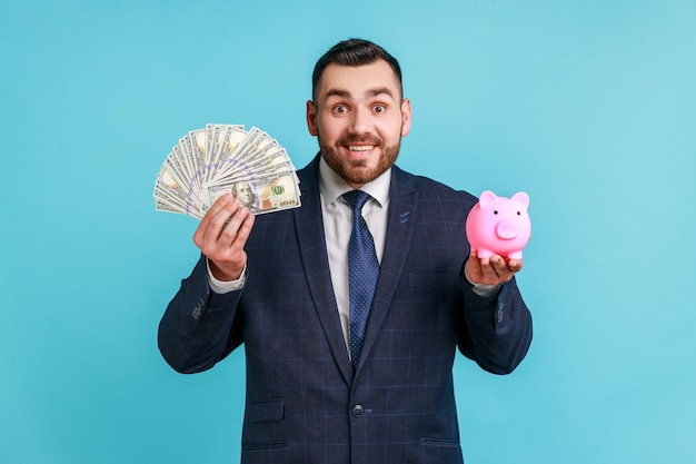 Smiling positive businessman with beard wearing official style suit holding dollar banknotes and piggy bank, savings, high rate deposit. Indoor studio shot isolated on blue background.