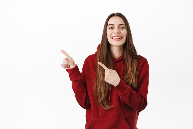 Smiling positive girl shows perfect white teeth grin pointing fingers aside at copy space promotional text on copy space give recommendation offer promo deal white background