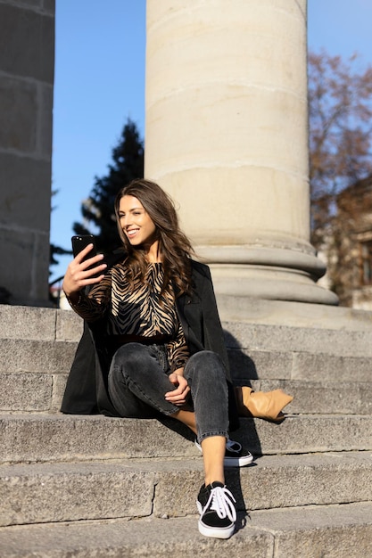 Smiling positive girl sitting on the stairs holding a phone shooting a video or taking a photo An influencer sits and talks to followers