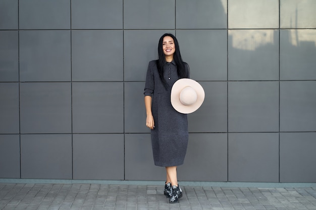 Smiling positive pretty young woman wearing youth grey oversize dress and fashionable shoes holding fashionable hat. Model in trendy outfit posing on urban grey wall. Outdoor portrait.