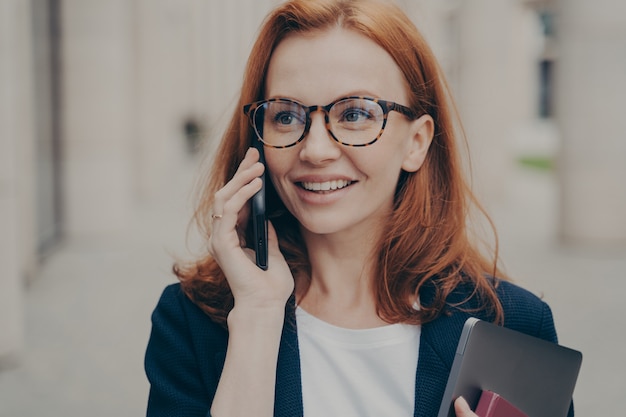 Smiling positive red-haired business woman wearing spectacles calling partner, standing outdoors