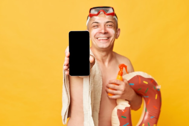 Smiling positive tourist man at summer resort standing with rubber ring isolated over yellow background showing cell phone with empty screen copy space for advertisement