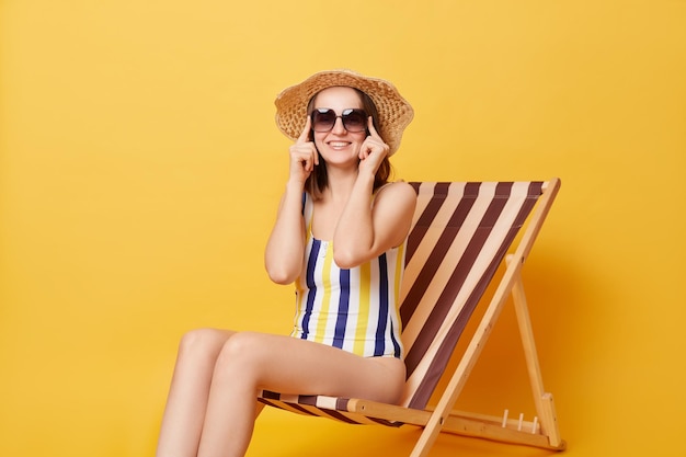 Smiling positive young woman wearing striped swimwear and hat sitting on deck chair isolated over yellow background holding her sunglasses looking at camera resting on resort