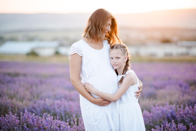 Smiling pregnant woman holding kid girl wearing white dresses posing in lavender flower meadow